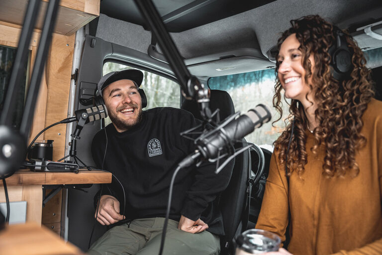 Adam Sauerwein and friend sitting in a van smiling with microphones in front of them. 