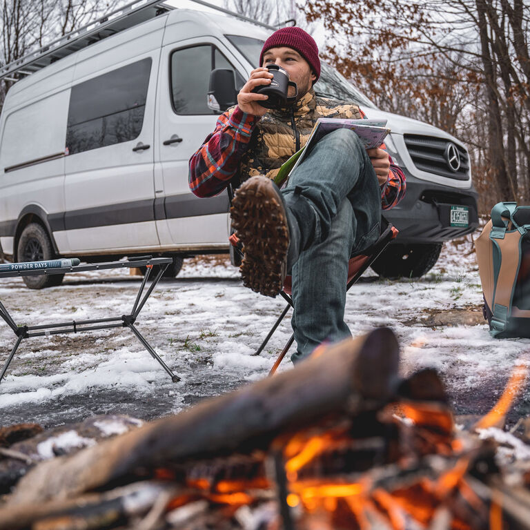 Adam Sauerwein sitting near a campfire drinking from a mug in a snowy forest with a sprinter van in the background.  