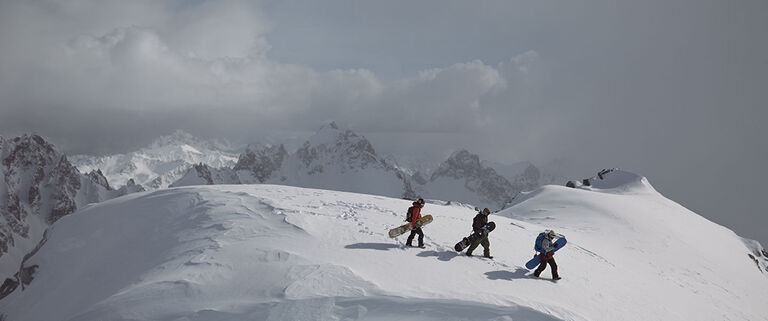 three snowboarders walking on a snow covered mountain summit with rocky snowy peaks in the background