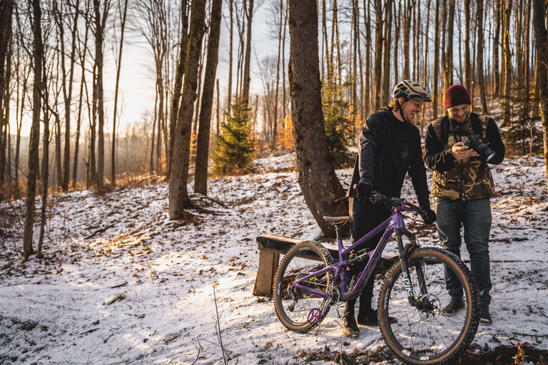 Adam Sauerwein and friend with a bike, laugh while looking at an image on a camera in a snowy forest area.