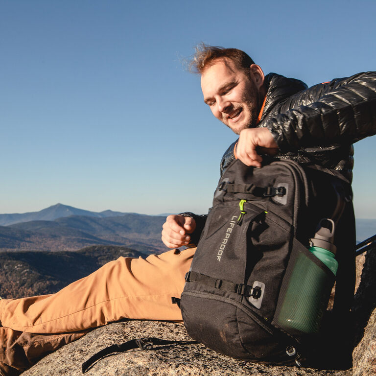Adam Sauerwein sitting on a rock holding his LifeProof backpack with mountains in the background.